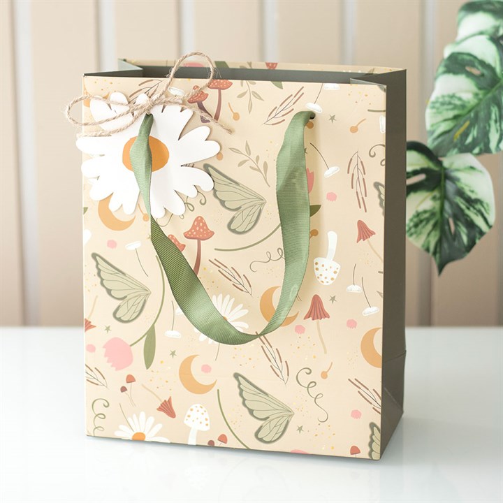 A pastel gift bag with green ribbon handles features whimsical illustrations of mushrooms, butterflies, flowers, leaves, and moons. Perfect for a magical present, it includes a white daisy tag and a leafy plant in the background.