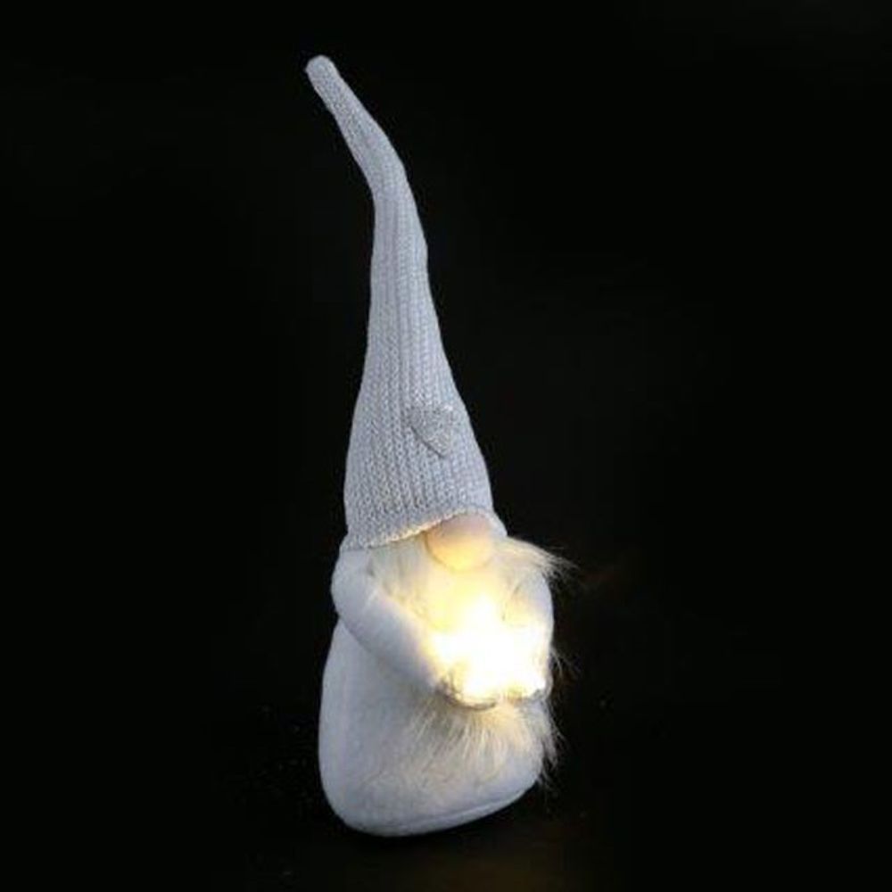 A small Christmas gnome decoration with a long, pointed white knitted hat and a fluffy beard holds a glowing, light-up star, set against a plain black background. The gnome’s face remains hidden.