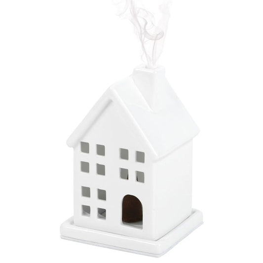 A white ceramic incense cone holder, shaped like a house with a pitched roof and square windows. Smoke gently rises from the chimney, fragrancing the air. The minimalist design sits smoothly on a flat base.