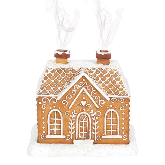 A gingerbread house with intricate white icing designs stands on a white base, serving as a festive incense cone burner. It features a textured roof, arched windows, and a detailed door. Two chimneys emit wispy streams of smoke against a snowy scene.