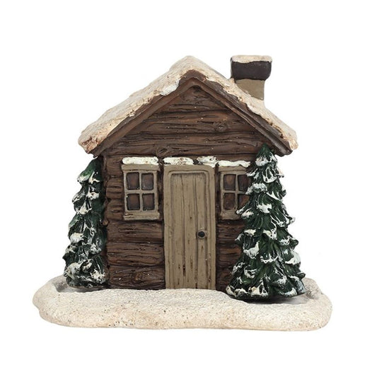 A cosy winter night scene unfolds with a small decorative log cabin, featuring a snow-covered roof and smoking chimney. Two snow-dusted evergreen trees flank the door, and textured snow blankets the base in wintry charm.