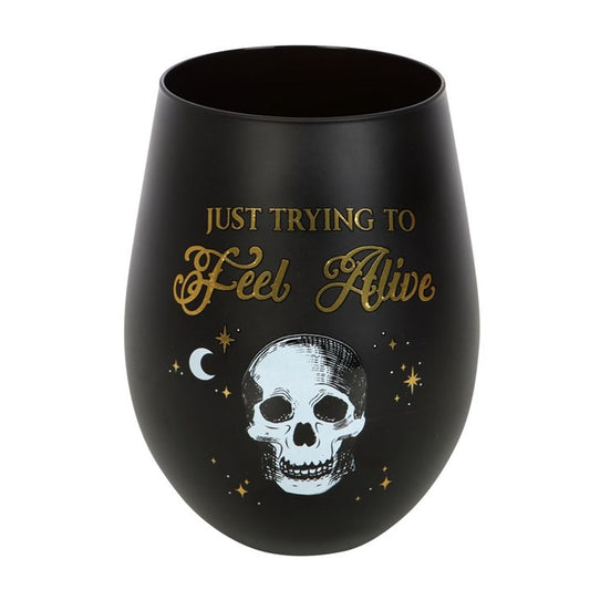 A matt black tumbler featuring gold and white text that reads Just trying to feel alive above a skeleton design. Embellished with small stars and a crescent moon, it exudes dark humour with its gothic and celestial vibe.