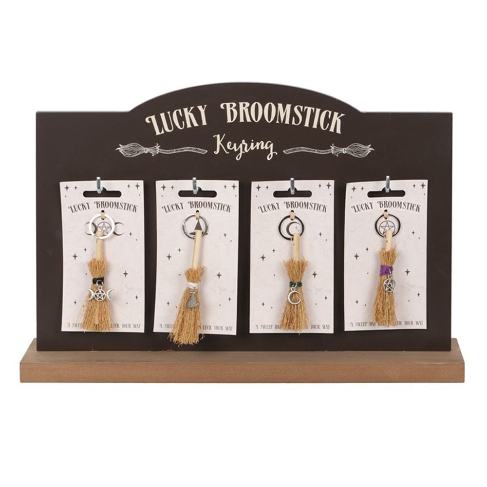 The display stand, labelled Lucky Broomstick Keyring, showcases magical designs with four enchanting keyrings. Each features a mini broomstick adorned with colourful threads and charms for luck and prosperity, elegantly hung on individual cards from a dark wooden base.
