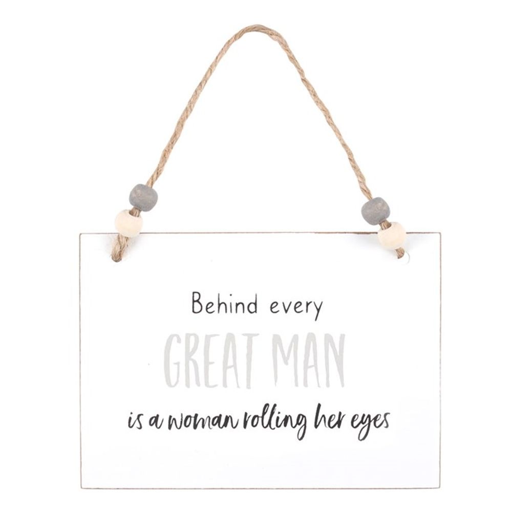 A white rectangular wooden hanging sign dangles from a twine hanger adorned with wooden beads. It humorously states, Behind every great man is a woman rolling her eyes, in mixed grey and black fonts, set against a plain background that highlights its witty charm—an ideal token gift.