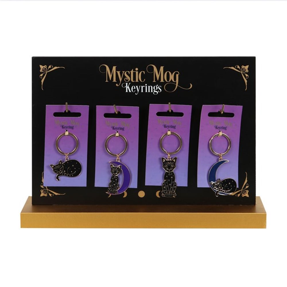 A counter display showcases four Mystic Mog keyrings on a sleek black and gold stand. Each black cat keyring, adorned with celestial design elements such as a sleeping cat and a cat sitting on a moon, is presented on a purple backing card against a plain white background.