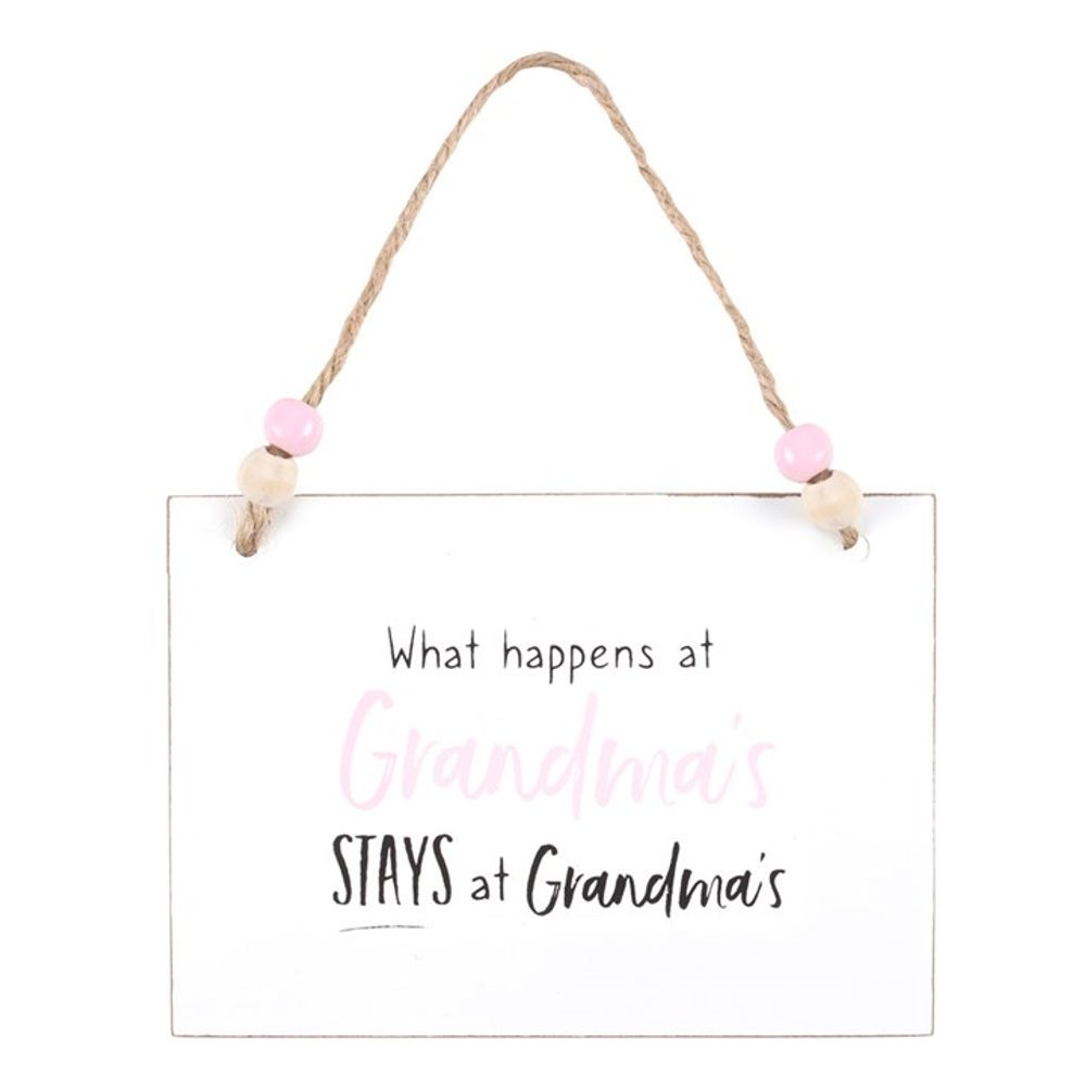 A rectangular white wooden hanging sign dangles from a jute string accented with pink and tan beads. Perfect as a gift for family and friends, the playful message reads, What happens at Grandmas STAYS at Grandmas, with Grandmas in cheerful pink against the simple design.