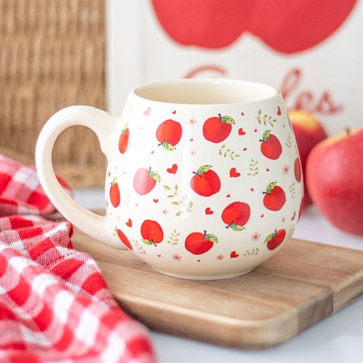 A cream-coloured ceramic mug with a rounded shape sits on a wooden board. The cosy cottagecore design features an apple motif, small hearts, and green leaves. A red apple and a chequered cloth complete the scene.