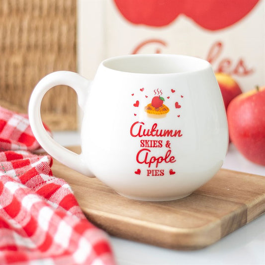 A ceramic mug with red text reading “Autumn Skies & Apple Pies”, decorated with hearts and a pie motif, brings cosy vibes as it sits on a wooden tray beside a red-checked cloth and a red apple.