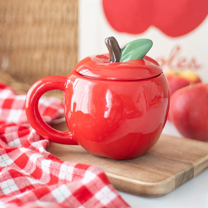This apple-shaped ceramic mug, one of our unique kitchen accessories, features a green leaf lid and rests on a wooden board beside a red-and-white chequered cloth and apple-themed décor.