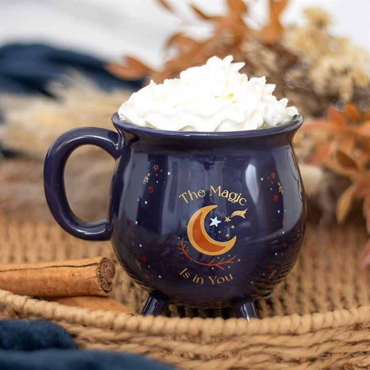 A mystical, dark blue cauldron-shaped mug filled with whipped cream rests on a woven surface. The words The Magic Is in You make it a perfect magical gift, with cinnamon sticks and dried flowers nearby.