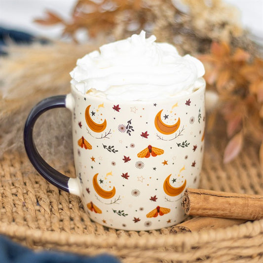 An ivory ceramic mug decorated with crescent moons, stars, and leaves contains an autumnal drink topped with whipped cream. A cinnamon stick rests nearby on a woven mat, set against a blurred harvest moon in the background.