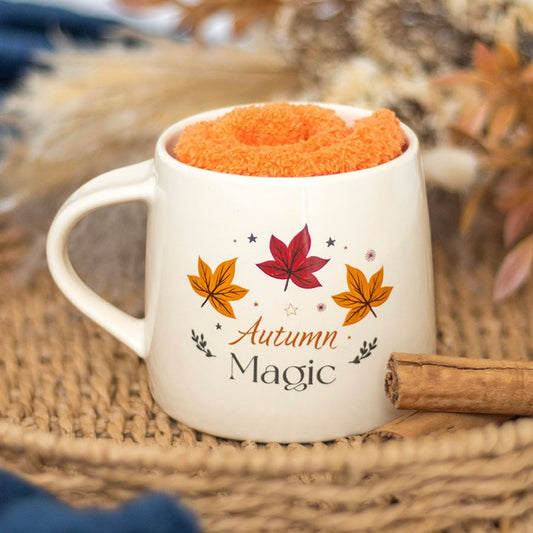 A ceramic mug bearing the words “Autumn Magic” and colourful leaves sits on a woven mat, filled with a soft orange cloth. A cinnamon stick lies beside it, with autumn-inspired socks and seasonal magic in the blurred background.
