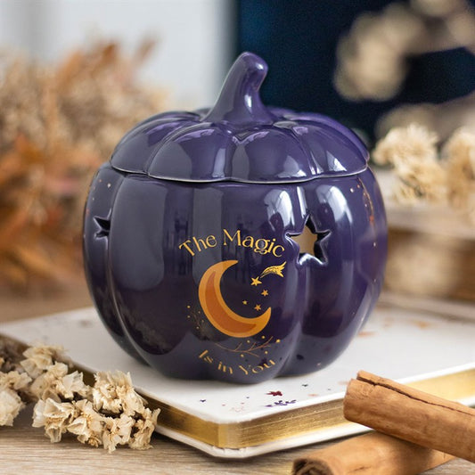 A glossy, pumpkin-shaped ceramic oil burner with star-shaped cut-outs and gold lettering reading “The Magic is in You” sits on a white surface beside dried flowers and cinnamon sticks.