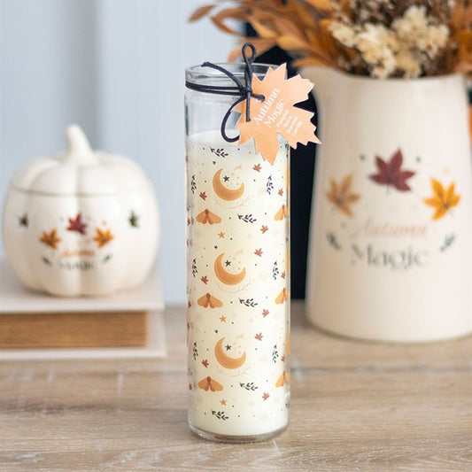 A tall glass tube candle decorated with orange leaves, crescent moons, and stars sits on a wooden surface. Perfect for autumn enthusiasts, it is paired with a pumpkin jar and an Autumn Magic vase of dried flowers.