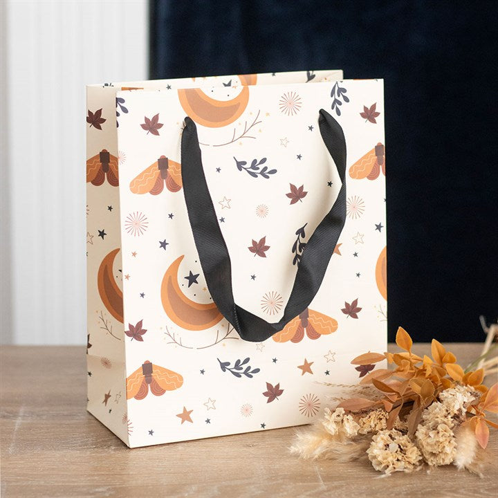 A decorative harvest moon-themed gift bag with black handles rests on a wooden surface. The bag features orange crescent moons, moths, and leaves on a cream background. Dried flowers are artfully arranged beside it, providing seasonal surprises.