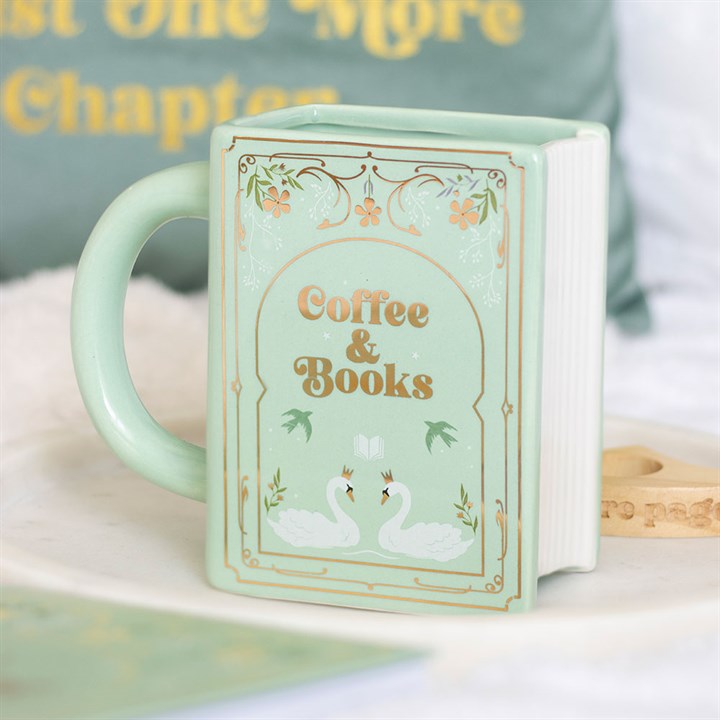 A pastel green ceramic coffee-and-books mug, shaped like a book with “Coffee & Books” and two white swans on the front, sits on a white surface. A blurred cushion and other objects appear in the background.
