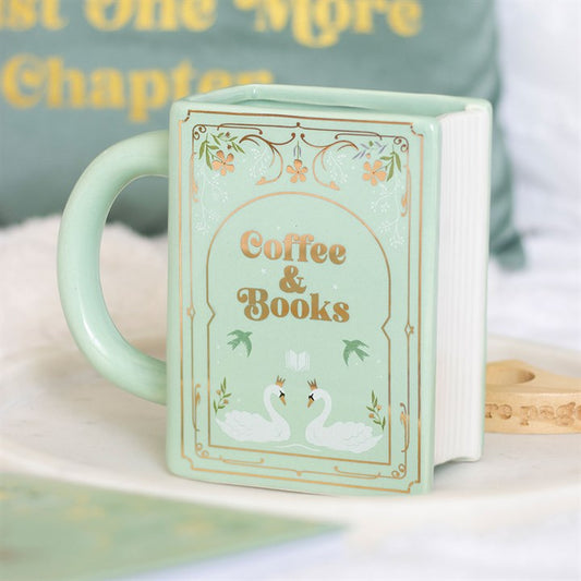 A pastel green ceramic coffee-and-books mug, shaped like a book with “Coffee & Books” and two white swans on the front, sits on a white surface. A blurred cushion and other objects appear in the background.