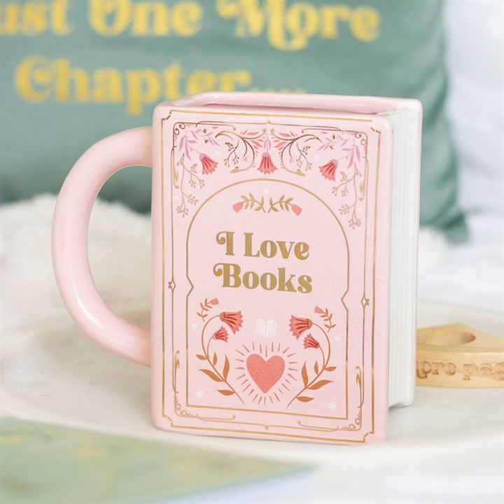A pink ceramic mug shaped like a book, featuring floral designs and the words “I Love Books” in gold, sits on a tray—a charming literary gift. In the background, a green cushion reads “Just One More Chapter.”.
