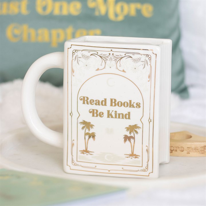 A cream-coloured, book-shaped mug features gold accents, two palm trees, and the words Read Books Be Kind. In the background, a cushion displays the phrase Just One More Chapter in yellow letters—a perfect gift for readers.
