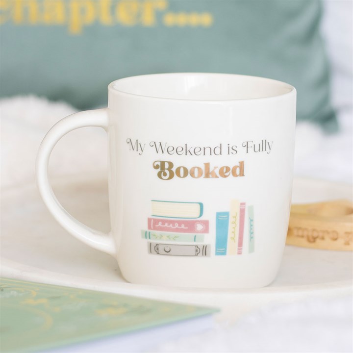 A ceramic mug featuring pastel-coloured books and the phrase “My Weekend is Fully Booked”—perfect for book lovers. Resting on a white tray beside a wooden bracelet, with a blurred book and cushion in the background.