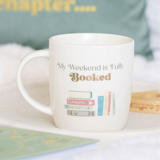 A ceramic mug featuring pastel-coloured books and the phrase “My Weekend is Fully Booked”—perfect for book lovers. Resting on a white tray beside a wooden bracelet, with a blurred book and cushion in the background.