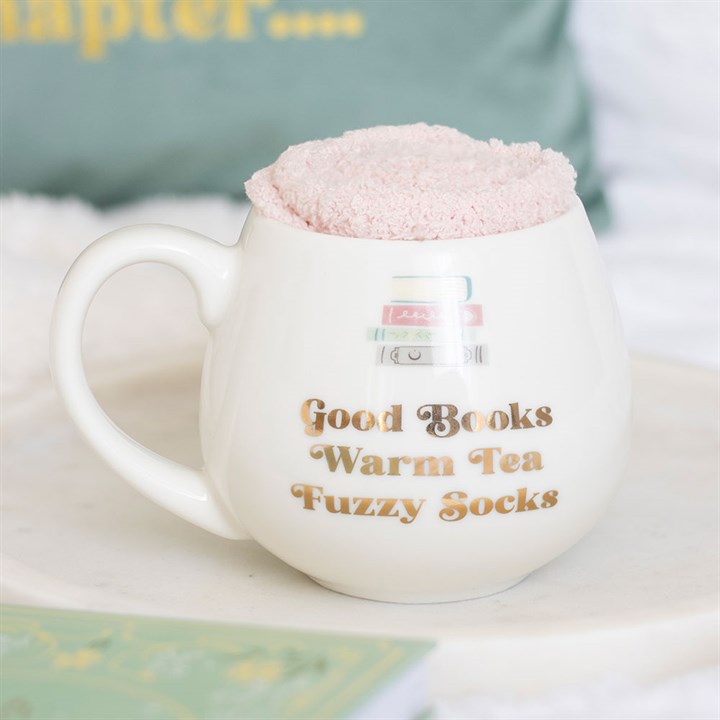 A ceramic mug with gold lettering reading ‘Good Books, Warm Tea, Fuzzy Socks’ sits on a white surface. Filled with pale pink fluffy socks, this sock gift set makes the perfect present for readers. A green cushion is blurred in the background.