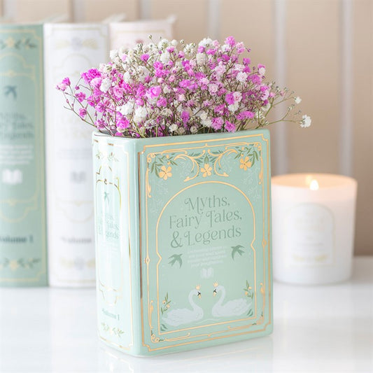 A mint-green book vase labelled “Myths, Fairy Tales & Legends” holds small pink and white flowers. Perfect home décor for book lovers, with a lit candle and similar books softly blurred in the background.