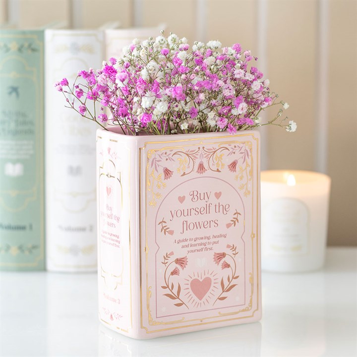 A pink ceramic book vase with gold accents and the text Buy yourself the flowers holds pink and white gypsophila. Pastel-coloured books and a lit white candle complete this charming home accessory for book lovers.