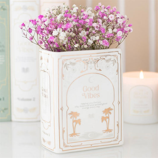 A white ceramic book-shaped vase with gold detailing and the text Good Vibes, perfect as a home accessory for book lovers, holds pink and white flowers. A lit candle and books in the background create a cosy atmosphere.