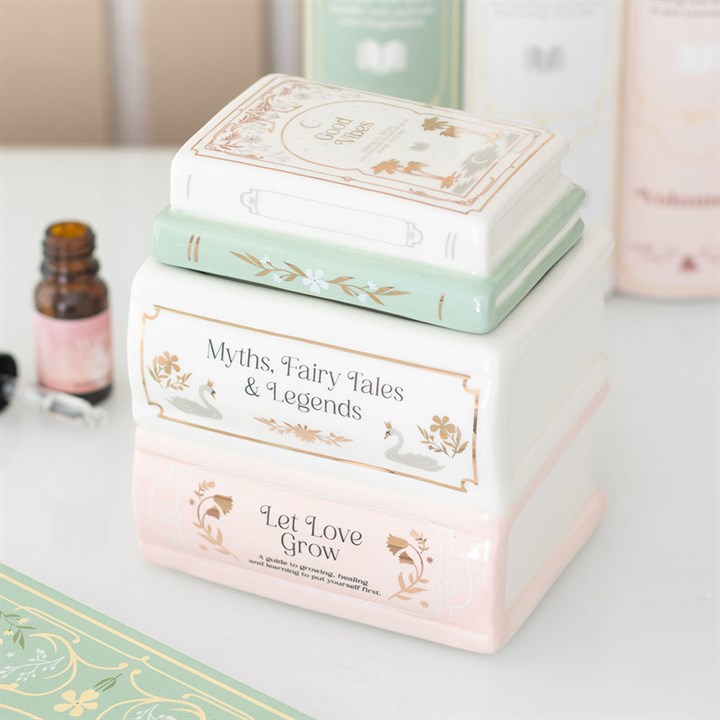 Three ceramic containers shaped like a stack of pastel books, each featuring decorative titles. A small brown dropper bottle rests nearby, ideal for use as a fragrance oil diffuser or ceramic oil burner.