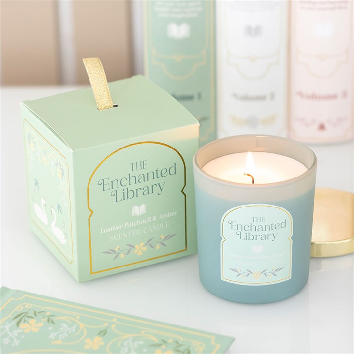 A lit fantasy-scented candle in a frosted jar labelled The Enchanted Library sits beside its mint-green box, perfect as a gift for book lovers. Pastel-coloured books and paper add charm to the cosy white setting.
