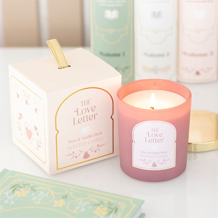 A pink rose, vanilla and musk candle labelled ‘The Love Letter’ burns beside its matching pastel box with gold accents. Perfect as a gift for book lovers, books and a gold lid sit in the cosy background.