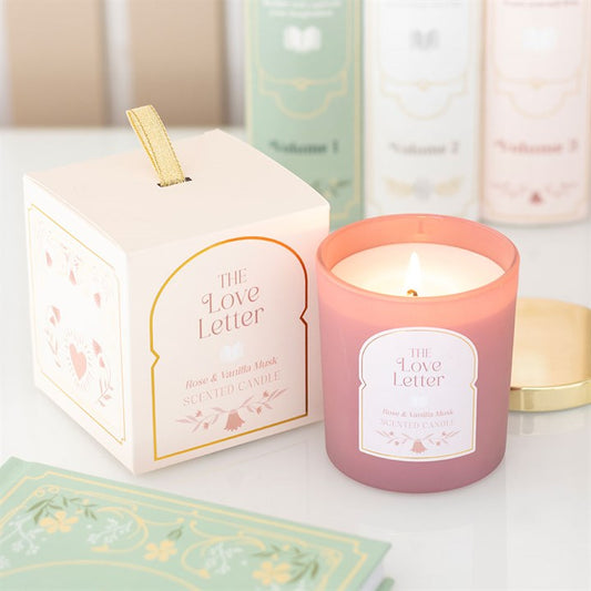 A pink rose, vanilla and musk candle labelled ‘The Love Letter’ burns beside its matching pastel box with gold accents. Perfect as a gift for book lovers, books and a gold lid sit in the cosy background.