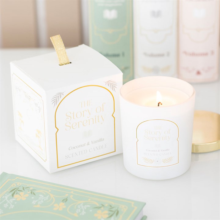 A white coconut and vanilla candle labelled The Story of Serenity burns beside its matching box—an inviting home fragrance and the perfect gift for book lovers. Soft pastel packaging and green envelopes are blurred in the background.