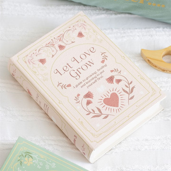 A pink and cream book titled Let Love Grow, featuring floral and heart illustrations, rests on white textured fabric—an ideal gift for book lovers. A partially visible mint-green book and a wooden teether are nearby.