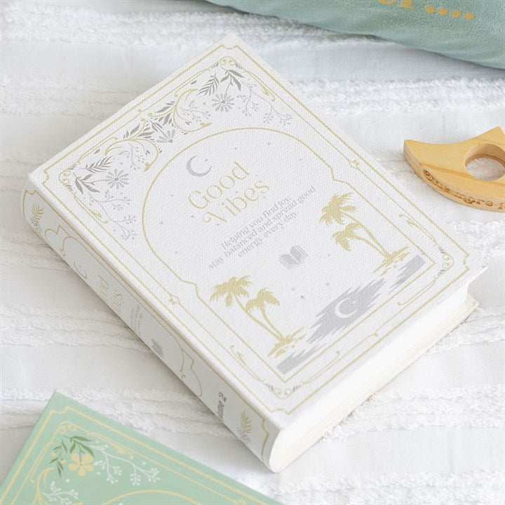 A white hardback book titled “Good Vibes”, featuring gold decorative designs and an eye-catching spine design, rests on a textured white surface next to a green book and a wooden ring toy.