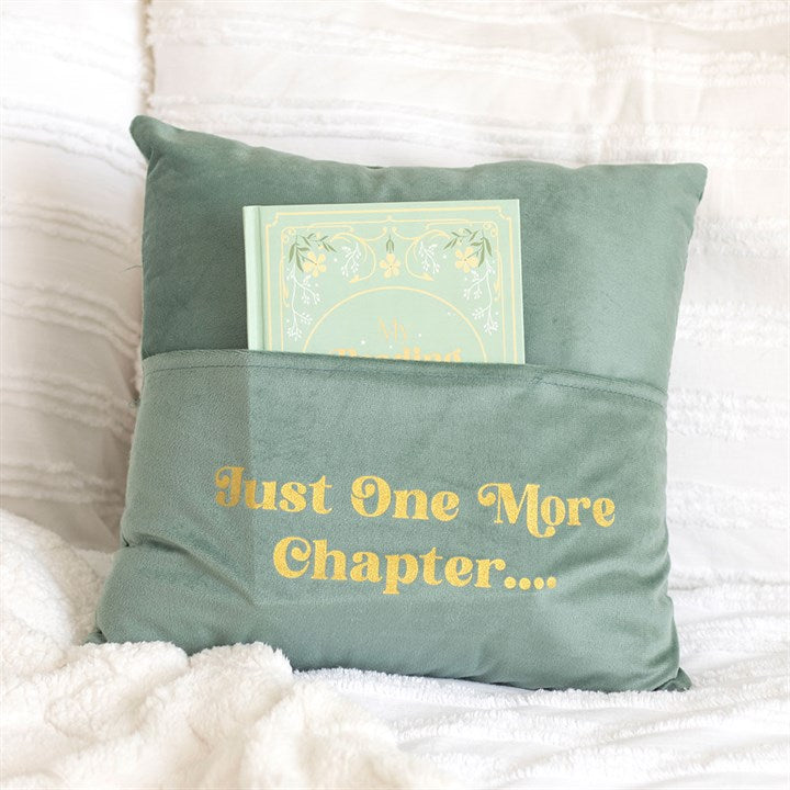 A soft green reading nook cushion with a front pocket holds a mint green book. Featuring yellow “Just One More Chapter…” text, it’s the perfect gift for book lovers to cosy up with on a white, textured blanket.