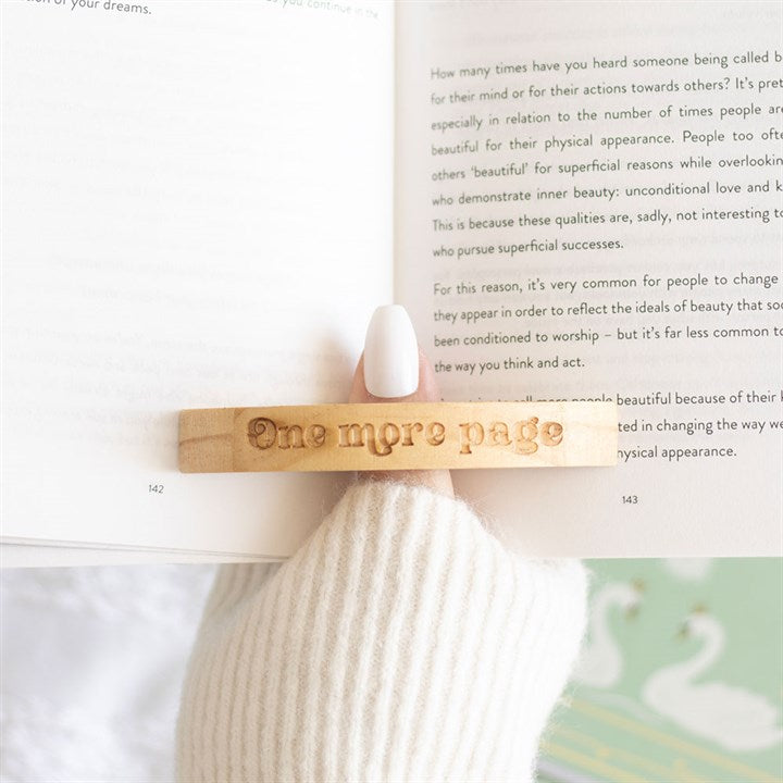 A hand with pale nail varnish uses a wooden thumb page holder engraved with “One more page” to keep a book open. The person is wearing a cream jumper; swan illustrations peek out from the blurred pages—a cosy scene for any bookworm.