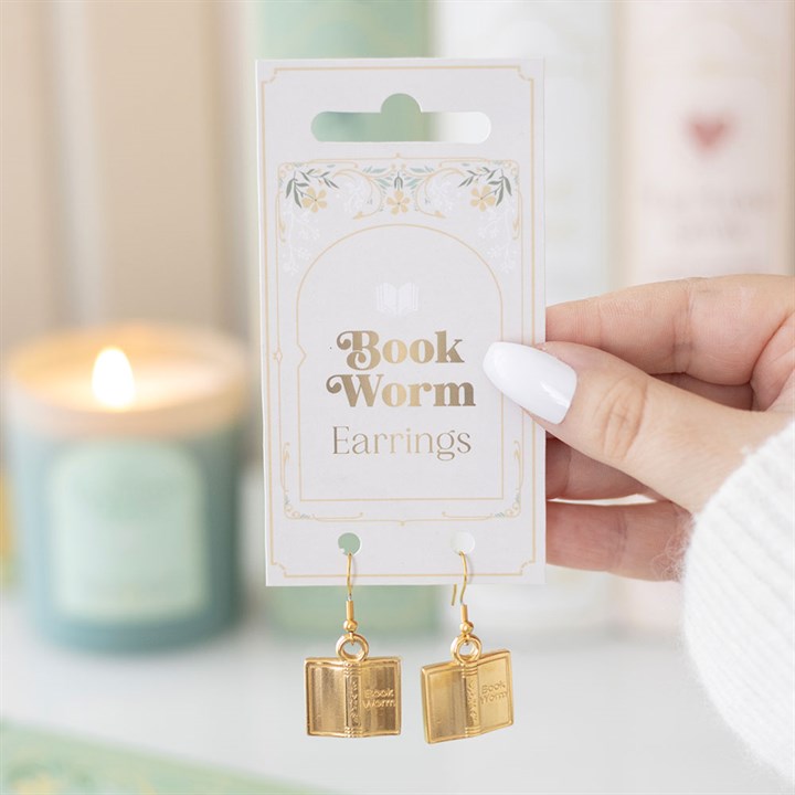 A hand with white-painted nails holds a card labelled Book Worm Earrings, displaying gold book-shaped earrings—an ideal gift for bibliophiles—with a lit candle and softly blurred pastel-coloured background elements.