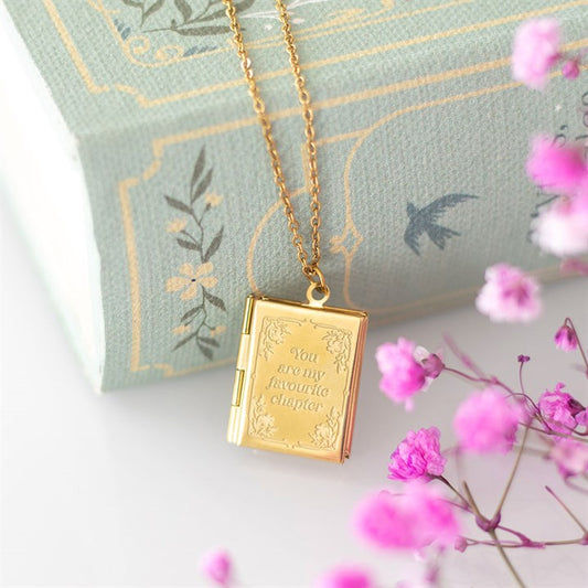 A gold, book-shaped locket necklace engraved with “You are my favourite chapter” rests on a pale green, floral book—an ideal literary-inspired gift for book lovers, accented by pink babys breath flowers.