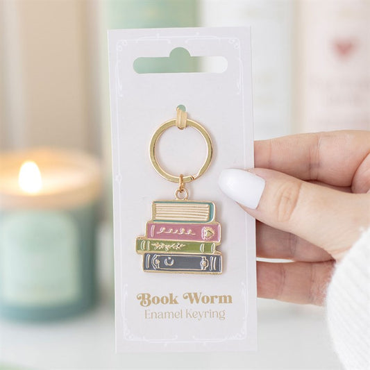 A hand with white nail varnish holds a gold Book Worm keyring—a charming reading accessory featuring a stack of colourful books, attached to a card labelled Book Worm Enamel Keyring. The perfect gift for bibliophiles.