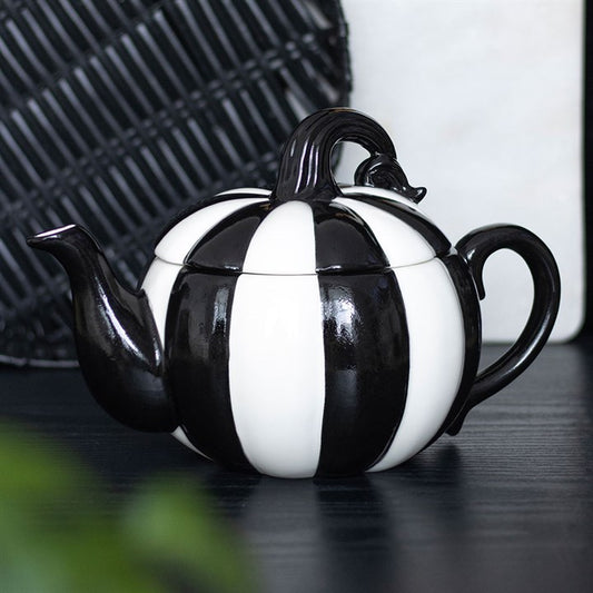 A black-and-white striped pumpkin teapot with a curving black stem sits on a dark surface—perfect for Halloween gatherings—beside a black woven mat against a white background.