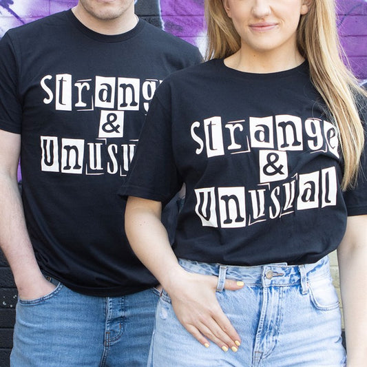 Two people stand side by side wearing unisex T-shirts emblazoned with the phrase “strange & unusual” in large, white, ransom-note-style lettering—a perfect look for spooky season against a wall painted in purple and black.