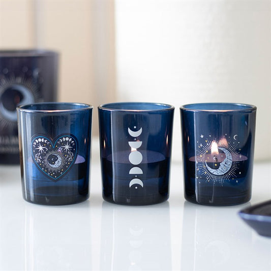 Three dark blue glass votive candle holders are positioned side by side. Each bears a white mystical motif: a heart with eyes and stars, moon phases in a row, and an all-seeing eye. One holder contains a lit candle.