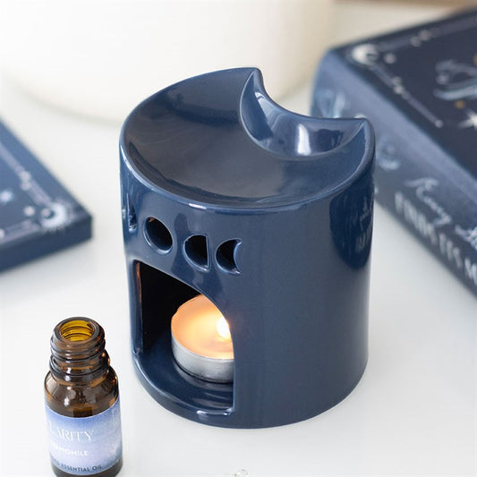 A blue crescent moon oil burner with circular cut-outs rests on a surface. A lit tealight glows within, with a bottle of chamomile essential oil nearby and books in the background.