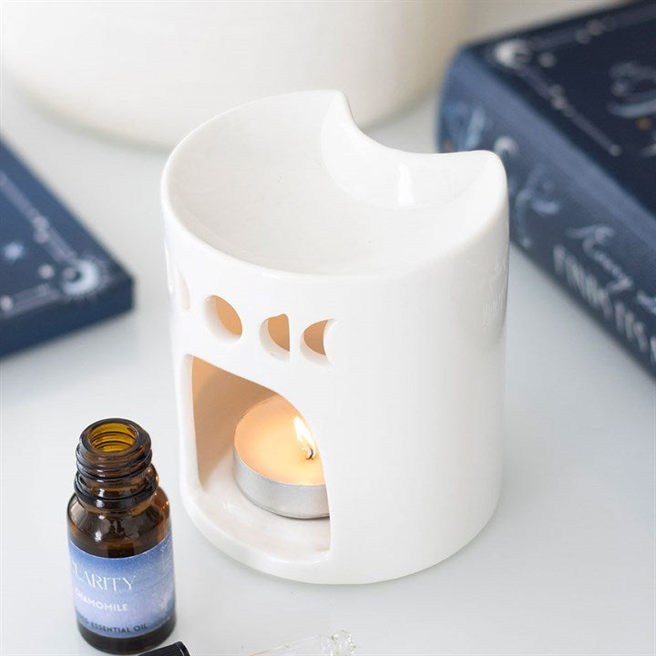 A white ceramic fragrance oil burner with round cut-outs and an open dish is placed beside a brown essential oil bottle and two dark blue books, lending celestial home décor charm to the scene.