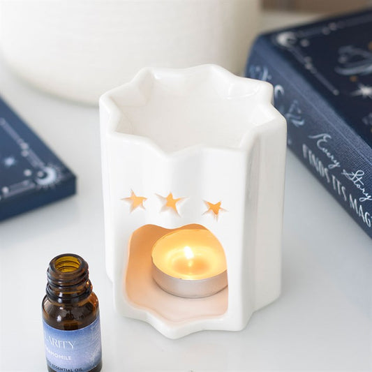 A star-shaped oil burner with wavy edges and three star cut-outs holds a glowing tealight inside. Fragrance oil, a blue book, and chamomile essential oil sit nearby on the white surface.