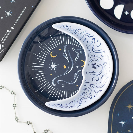 A round navy stacking trinket dish displays a painted moon with a face and swirling blue designs, set against a starry sky with celestial motifs. Nearby lie star-themed items and a silver star necklace.