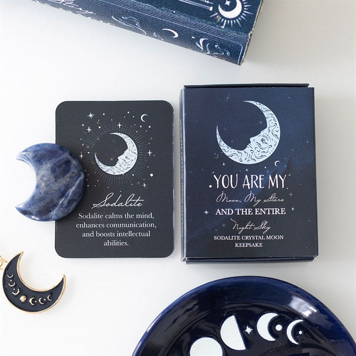 A card and box featuring celestial moon designs read, Sodalite calms the mind... Ideal for crystal collectors or moon rituals, a sodalite moon crystal and dark plate with crescent cut-outs are nearby.