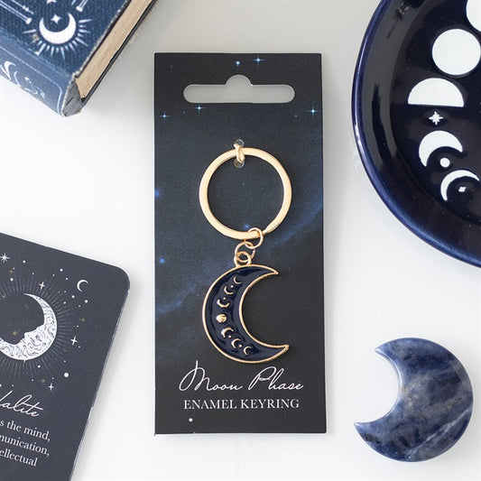 A celestial accessory, this crescent moon keyring with gold accents is attached to a black Moon Phase Enamel Keyring card. Nearby are a moon phase card, a blue dish with moon motifs, a blue crystal, and part of a book.